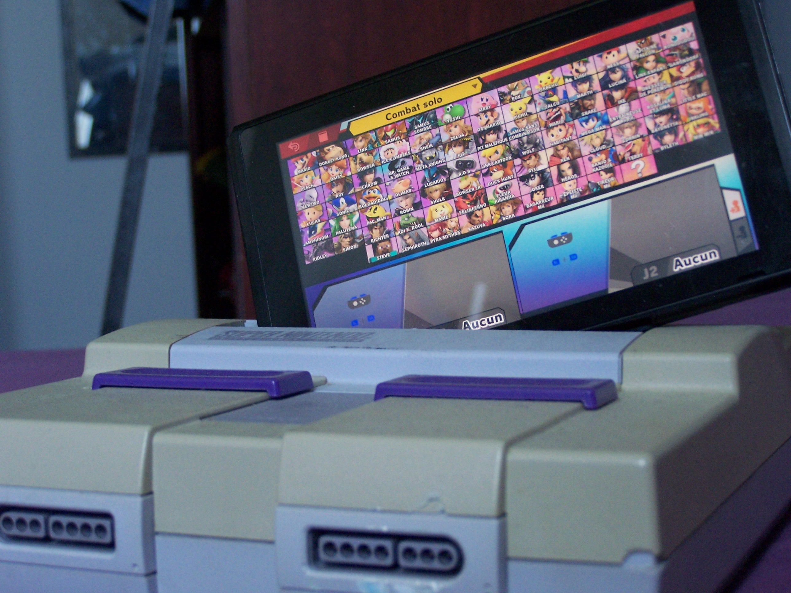 handheld game console with a fighting game character select screen showing, sat lopsided in the cartridge slot for a home game console.