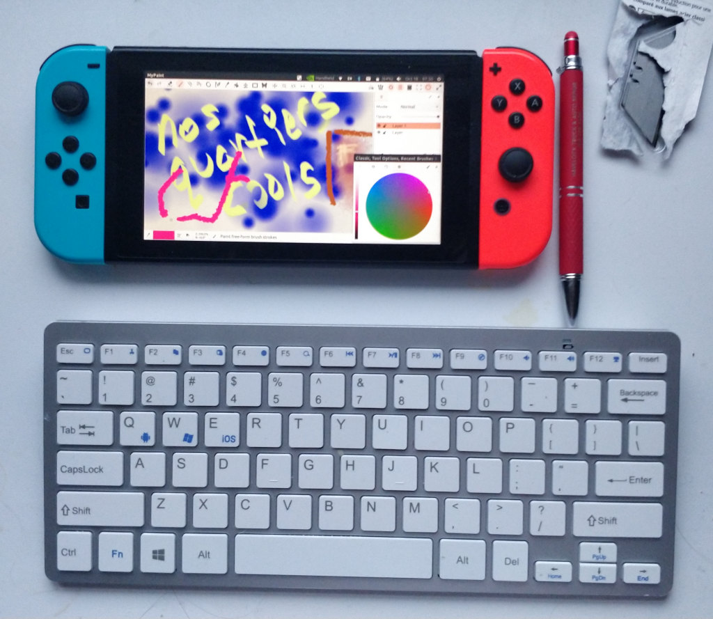 handheld gaming console with a drawing program open, a pen with rubber end, and a wireless keyboard, all sitting on a white desk.