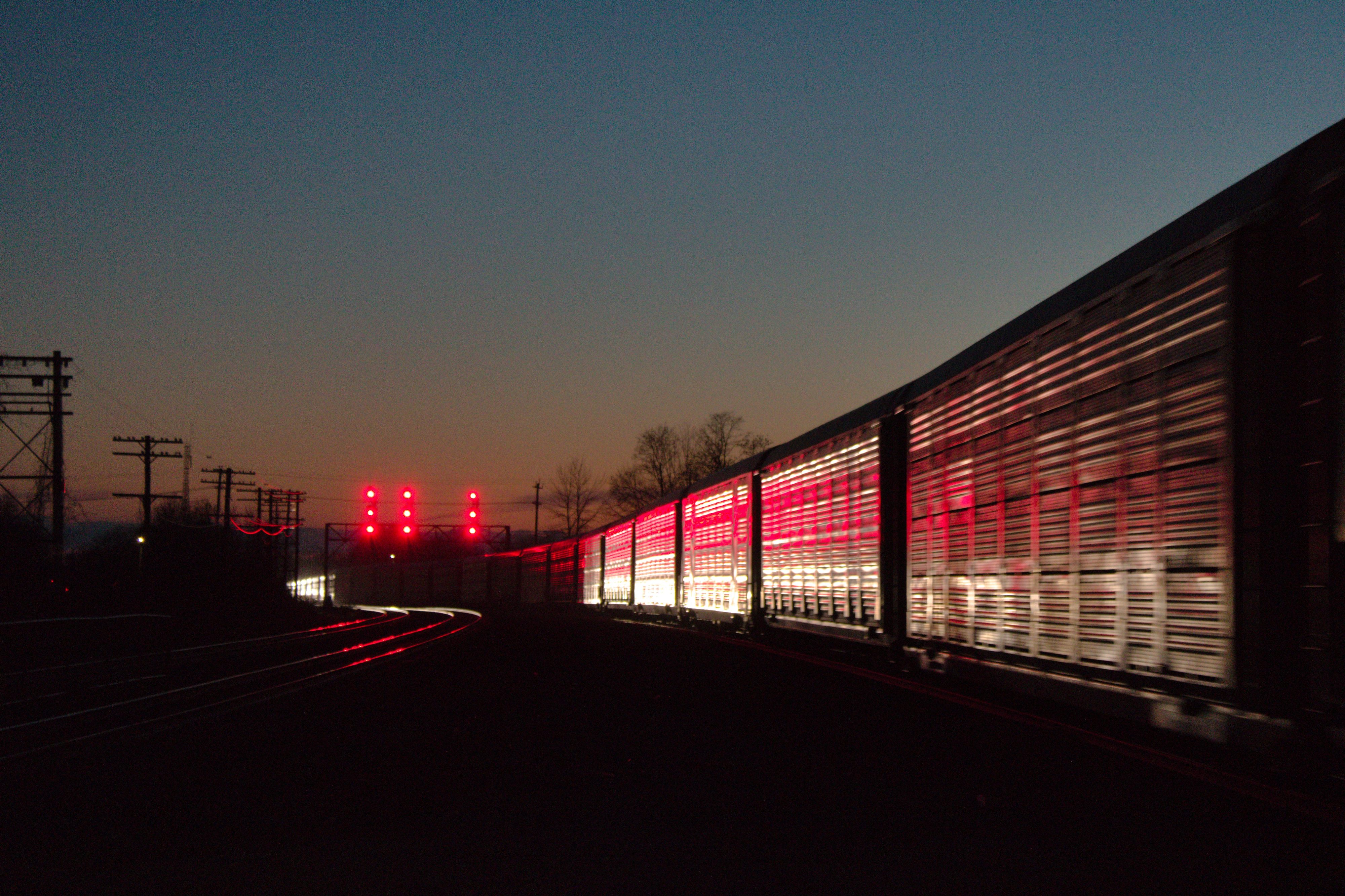 night photograph between railtracks with a signal bridge in the distance. there is a silver train car that the signals are reflecting off of dramatically. the ground and background are shadowed.