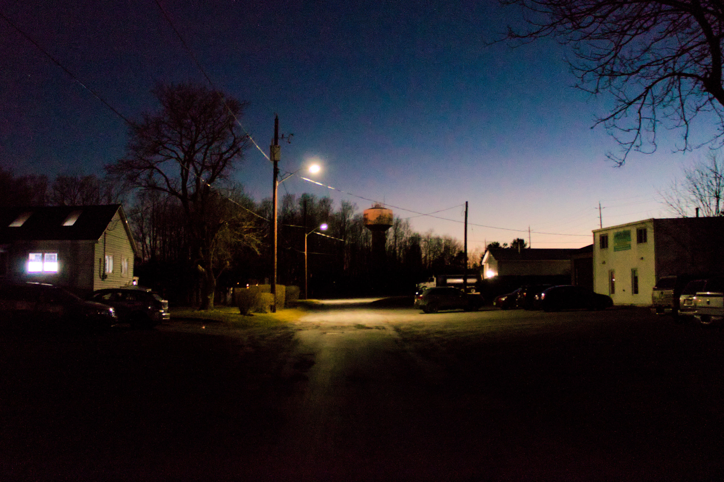 wide angle photo on a semirural industrial street with a forest at the end at night time. the street lamp contrasts heavily with the darkness, and there is some coloured noise in the dark blue sky.