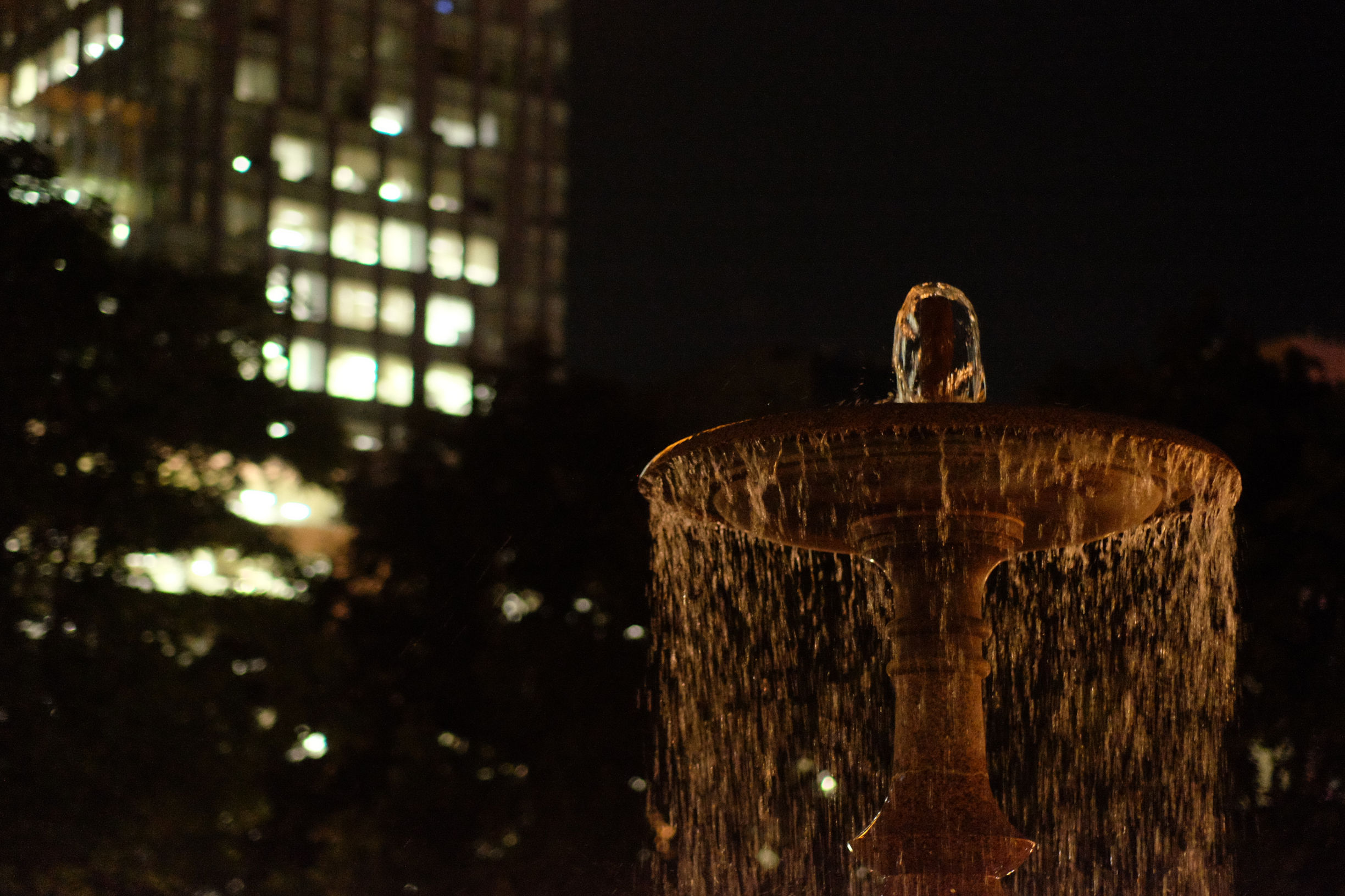water fountain at night time with a high rise building with lights on in the background.