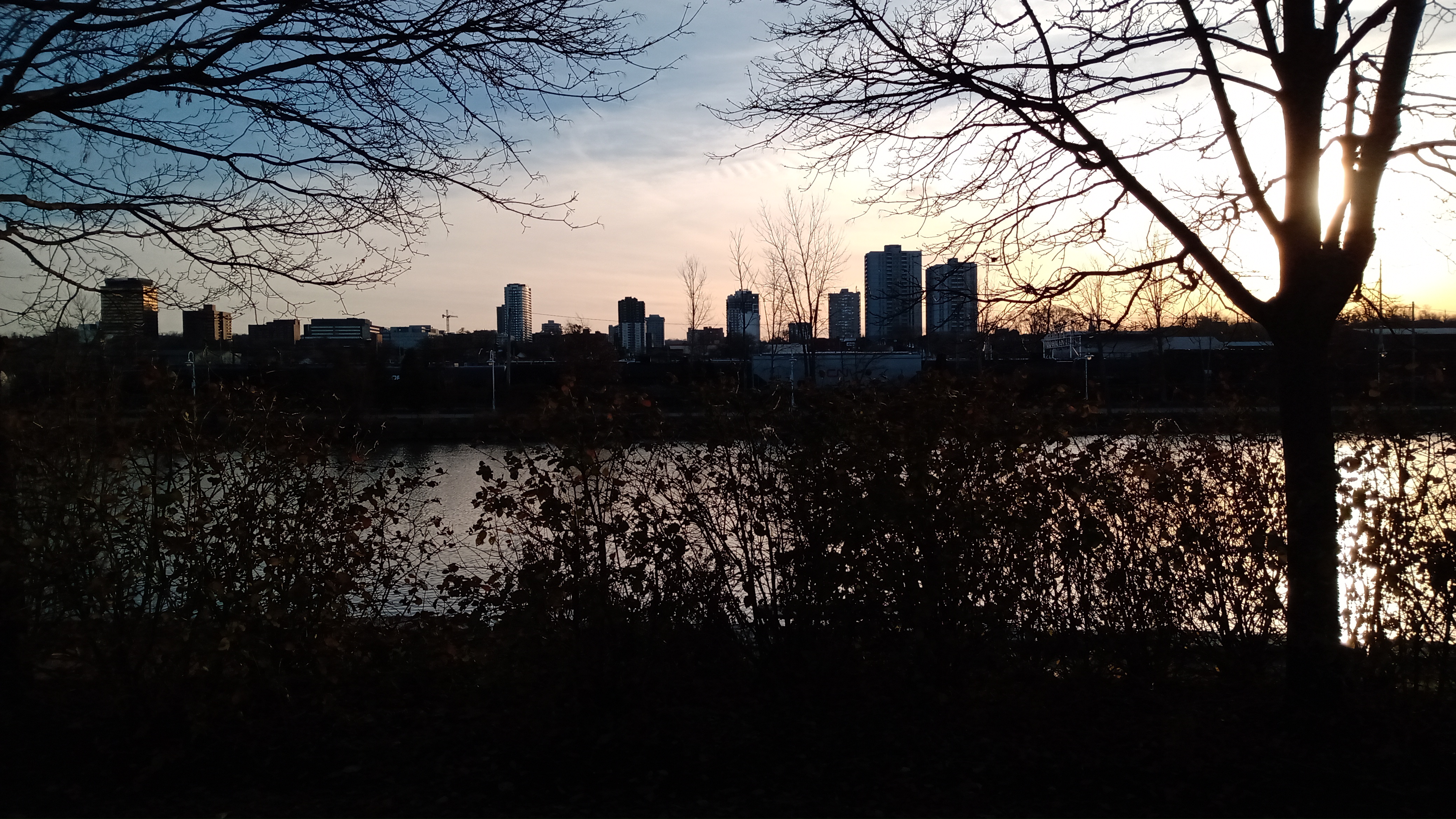 winter sunset, with bright orange on the right sky and a foggy dark blue on the left sky. the foreground has the silhouettes of trees, and then a water body, and then a city skyline.