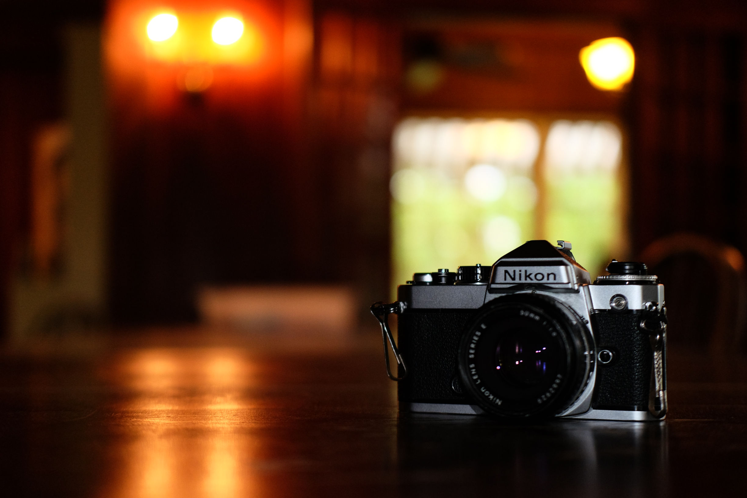 silver nikon film camera on a wooden table in a dark orange room. there is a single out of focus orange light source in the background.