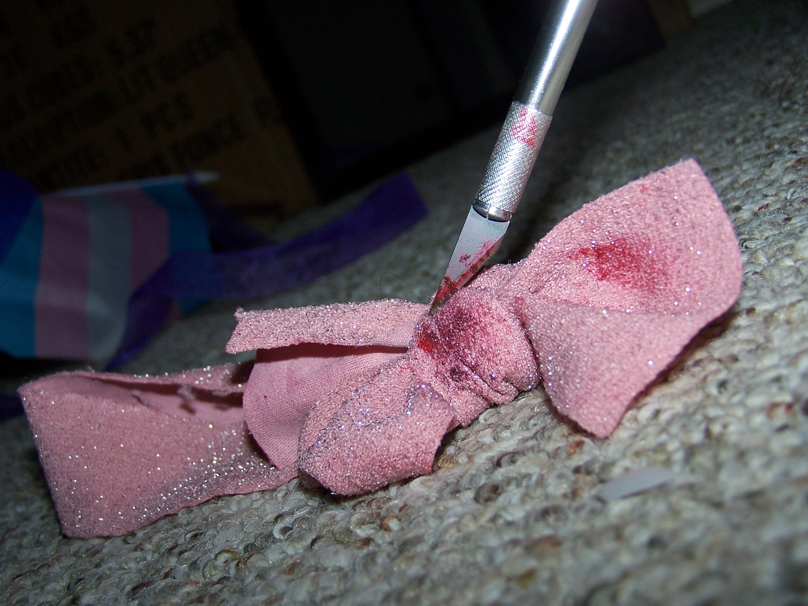 pink ribbon laying on a carpeted floor with a razor attached to a handle slanted into it from above. Both are stained red. The razor's steel and the sparkles in the ribbon are very bright. The background is nearly entirely dark and blurry.