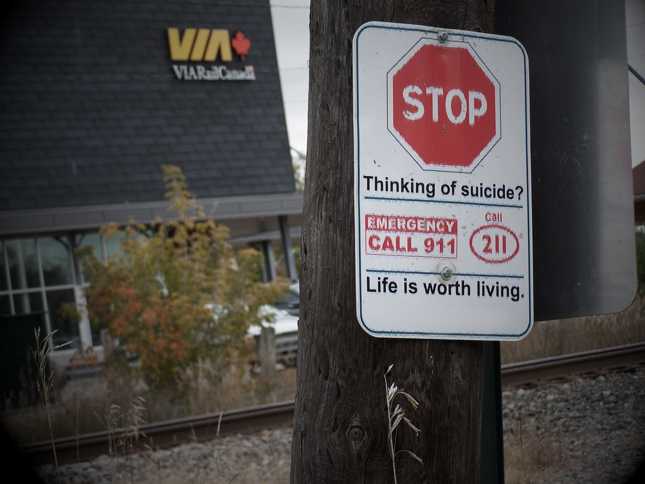 stop sign on a pole in front of a train track and a train station. under the stop sign says 'thinking of suicide?'.