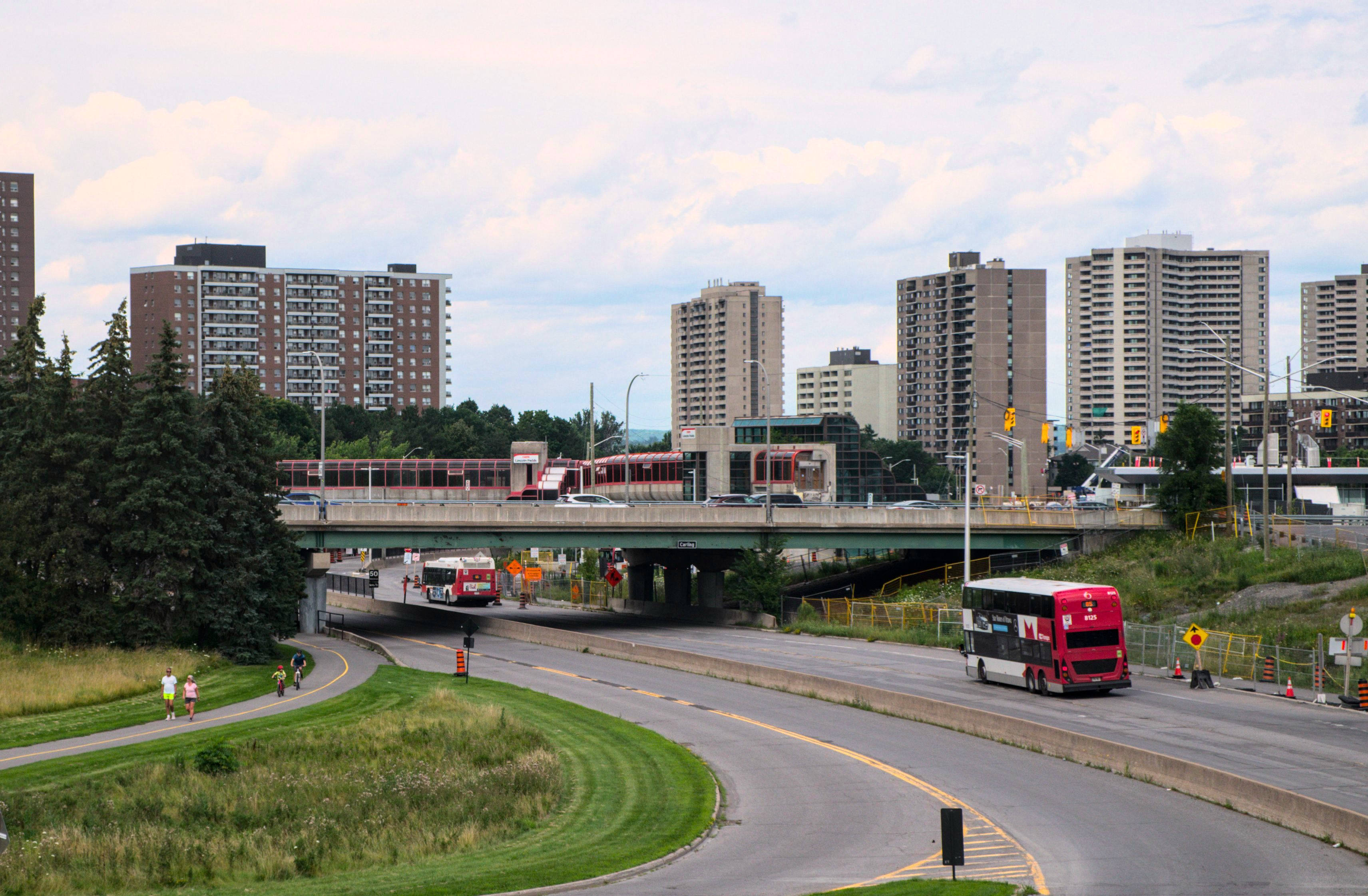 red double decker bus passing underneath a bridge towards a station, with apartment towers in the background.