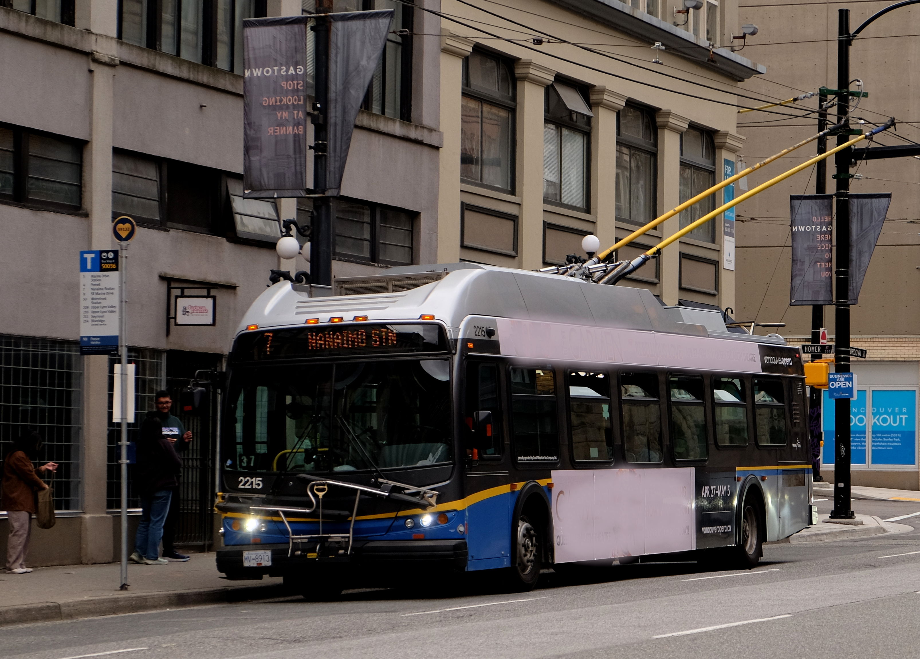trolleybus at a bus stop in front of a grey building.