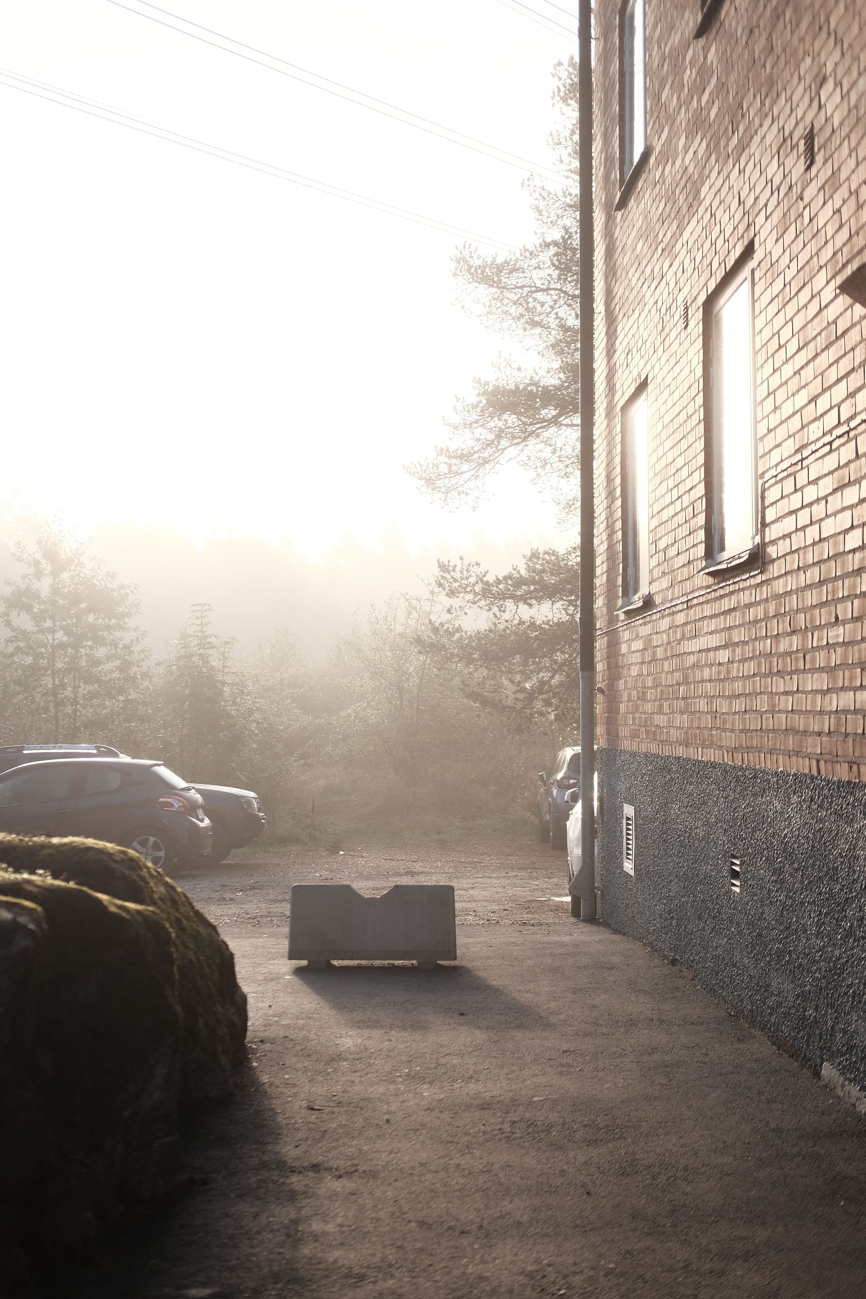 asphalt walkway towards a apartment building car park, with the building on the right and foliage in the background. the sky is brightly lit, but shrouded in thick fog.