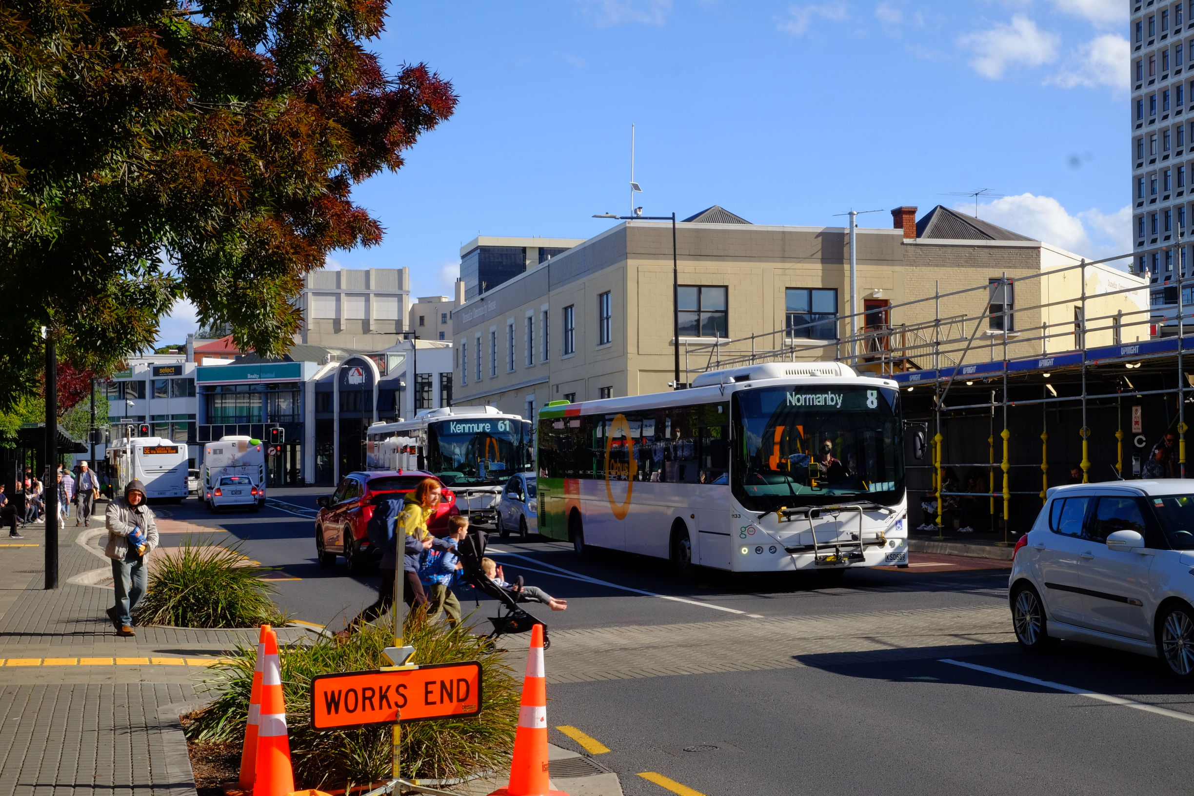woman with a pram crossing a street with buses and cars.