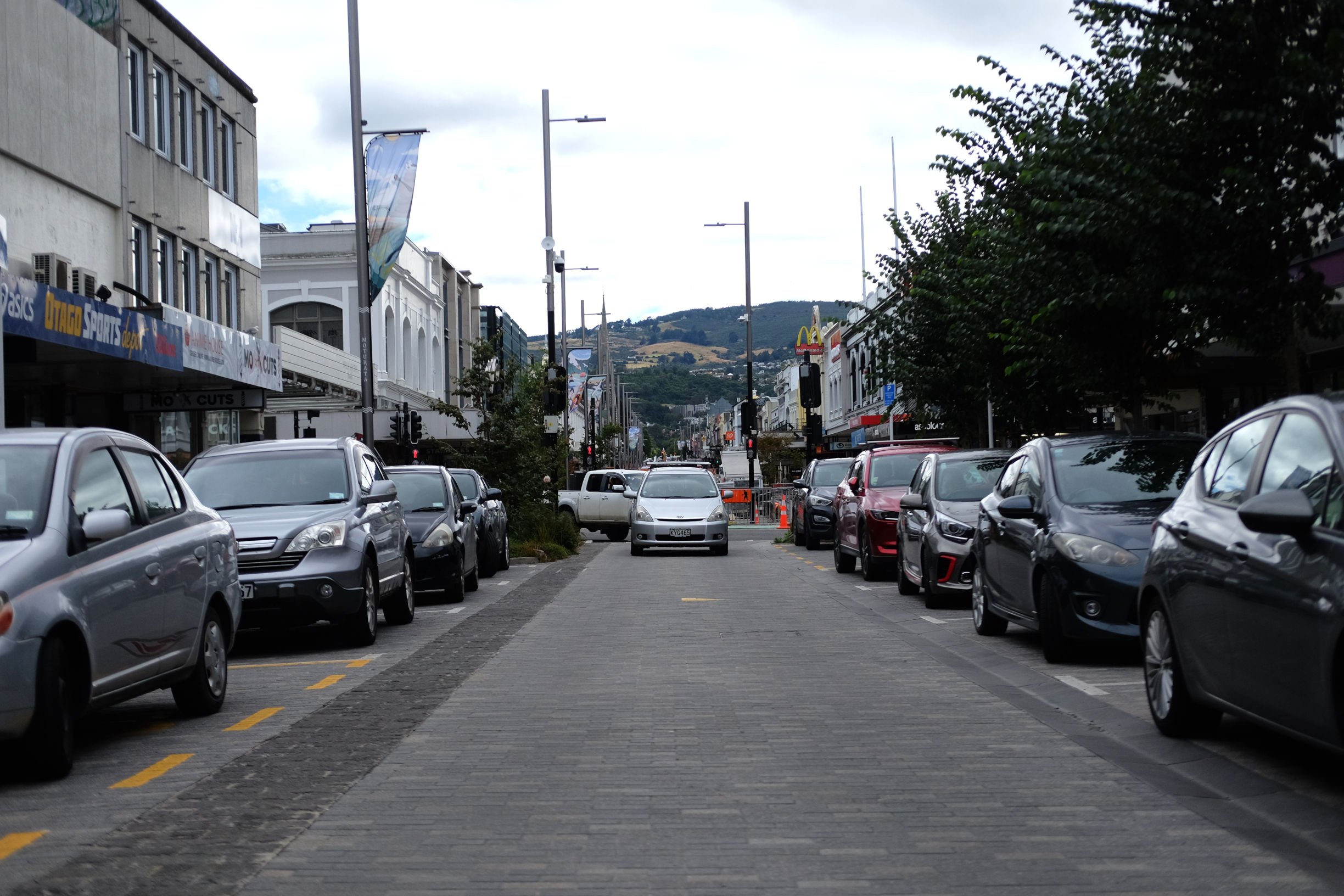 street with grey paving stones lined with parked cars, and a automobile oncoming.
