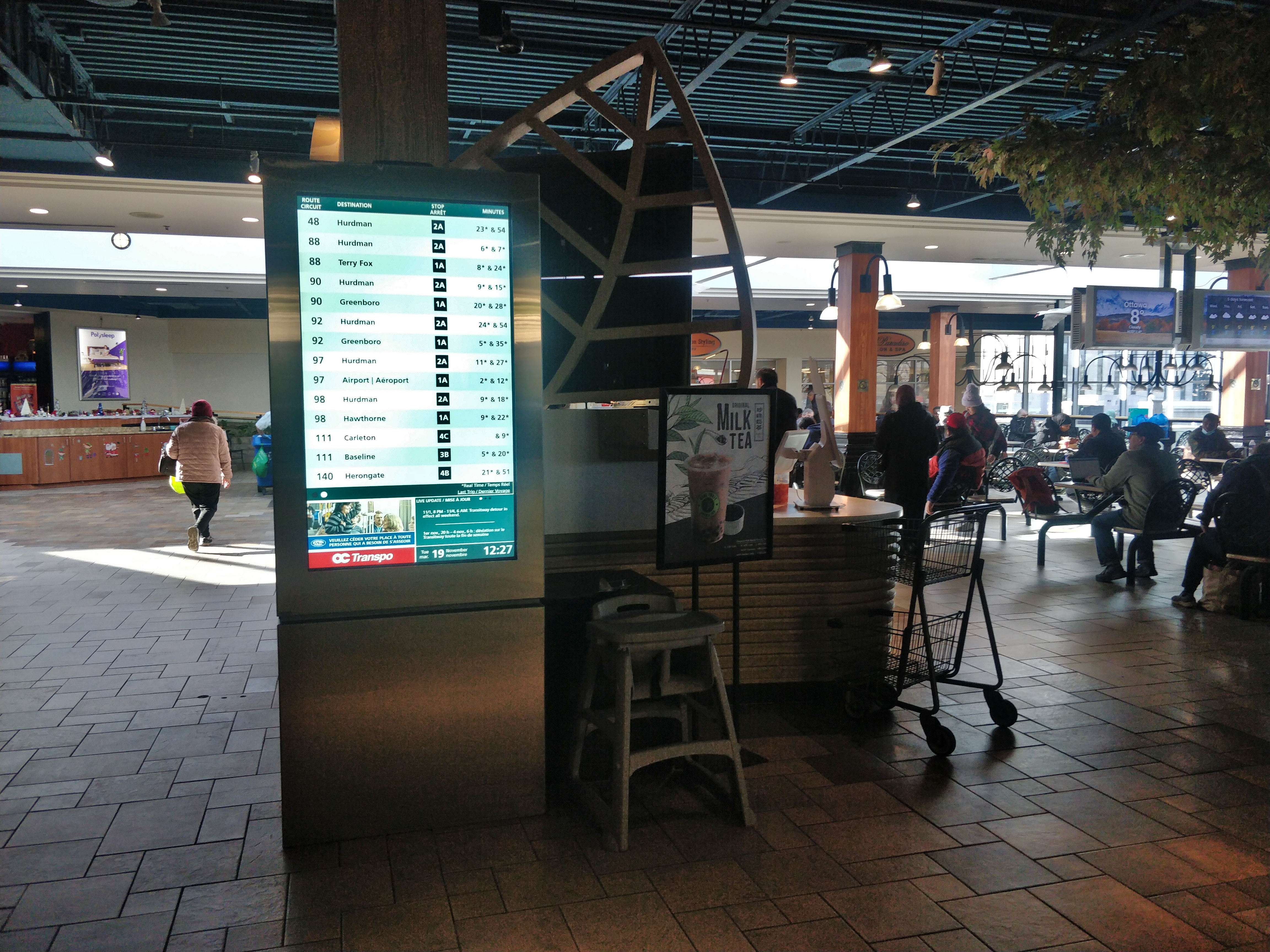 Bright blue tall screen with many bus routes on a tuesday at noon, in a mall food court.