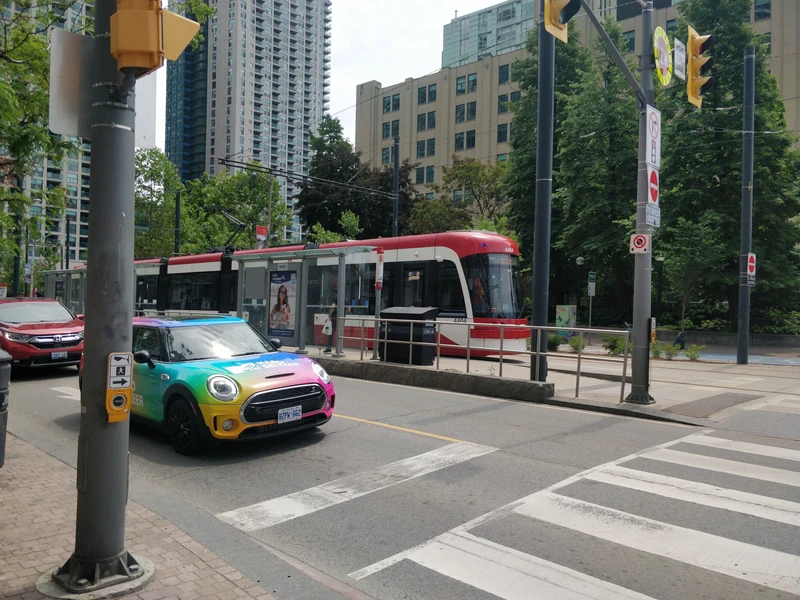 red tram with white strips along its length in its own right of way next to small roadway. it is travelling through a area of mixed mid and high rise buildings, with various architecture and construction materials. it is sunny, and there are lots of mature trees.