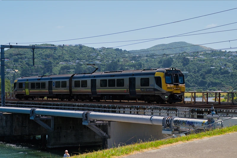 two car matangi trainset with driving ends on both sides and a articulating section in the midle. it is on a bridge over a large river in front of grassy steep hills. the side of the vehicle is grey, with the front face being yellow. it has visible hand holds on the exterior, and is vaguely barrel shaped.