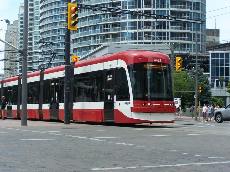 photograph of a tram in a blue glass condo district. pretty asphalt, red tram with white accents on border of the windows. Modern low floor design.