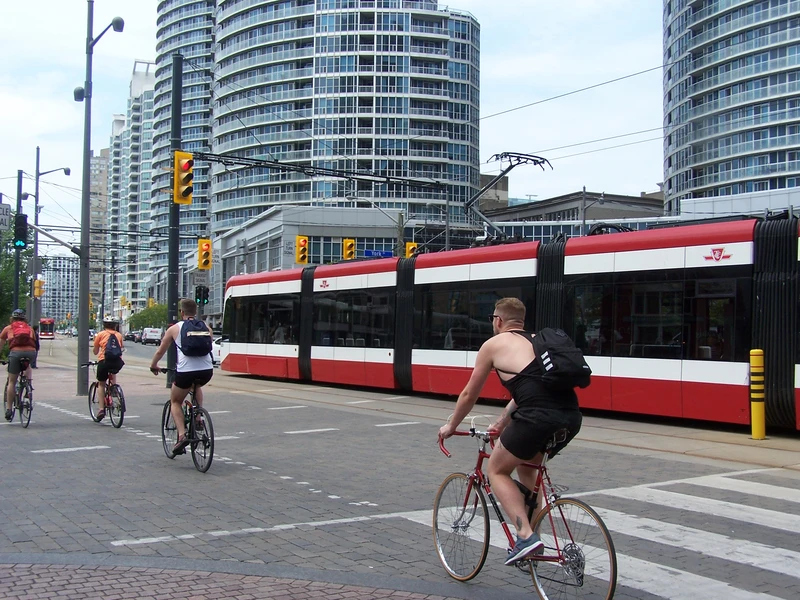 red tram with white stripes travelling in front of several blue and white glass towers. in the foreground, there are lots of cyclists travelling in the same direction as the tram.